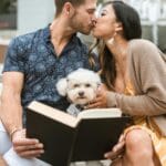 Para całuje się nad książką przy fontannie, obok siedzi biały pudel